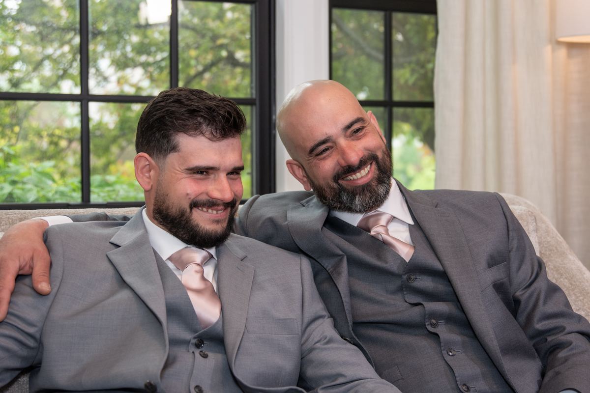 Two men in grey suits and pink ties smile warmly at each other on a sofa, illuminated by soft natural light from windows.
