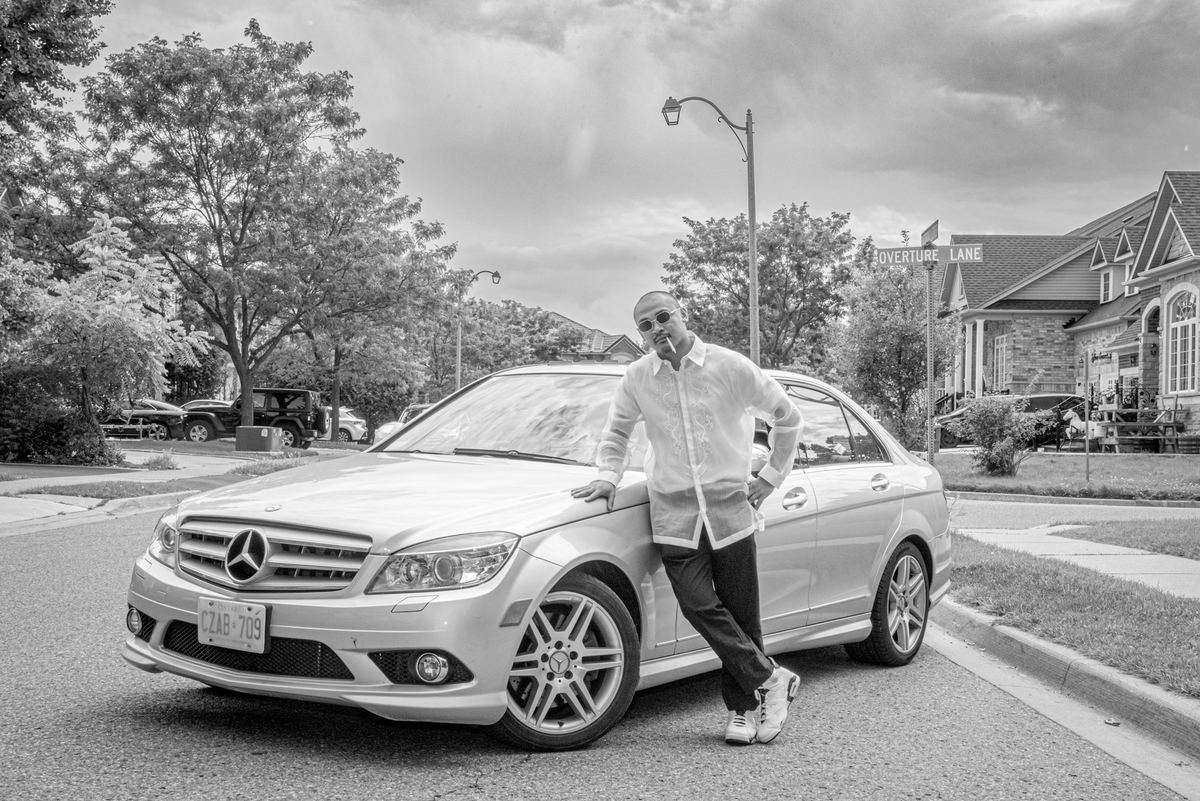 A man in a white shirt and sunglasses leans against a silver car on an overcast suburban street, emphasizing shadows.