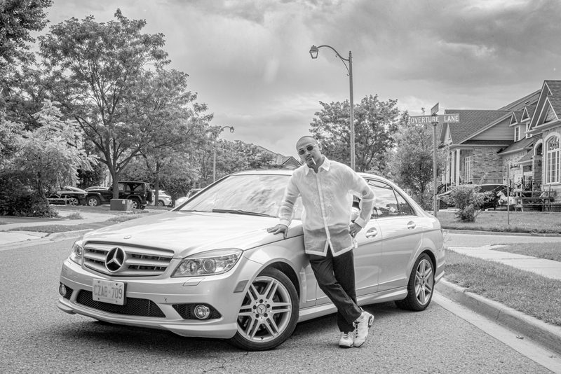 A man in a white shirt and sunglasses leans against a silver car on an overcast suburban street, emphasizing shadows.