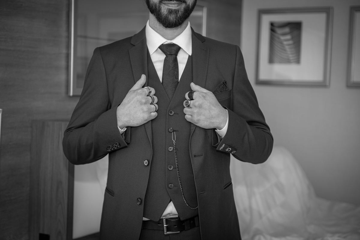 A groom adjusts his three-piece suit, smiling confidently in soft indoor lighting, fingers resting on rings.