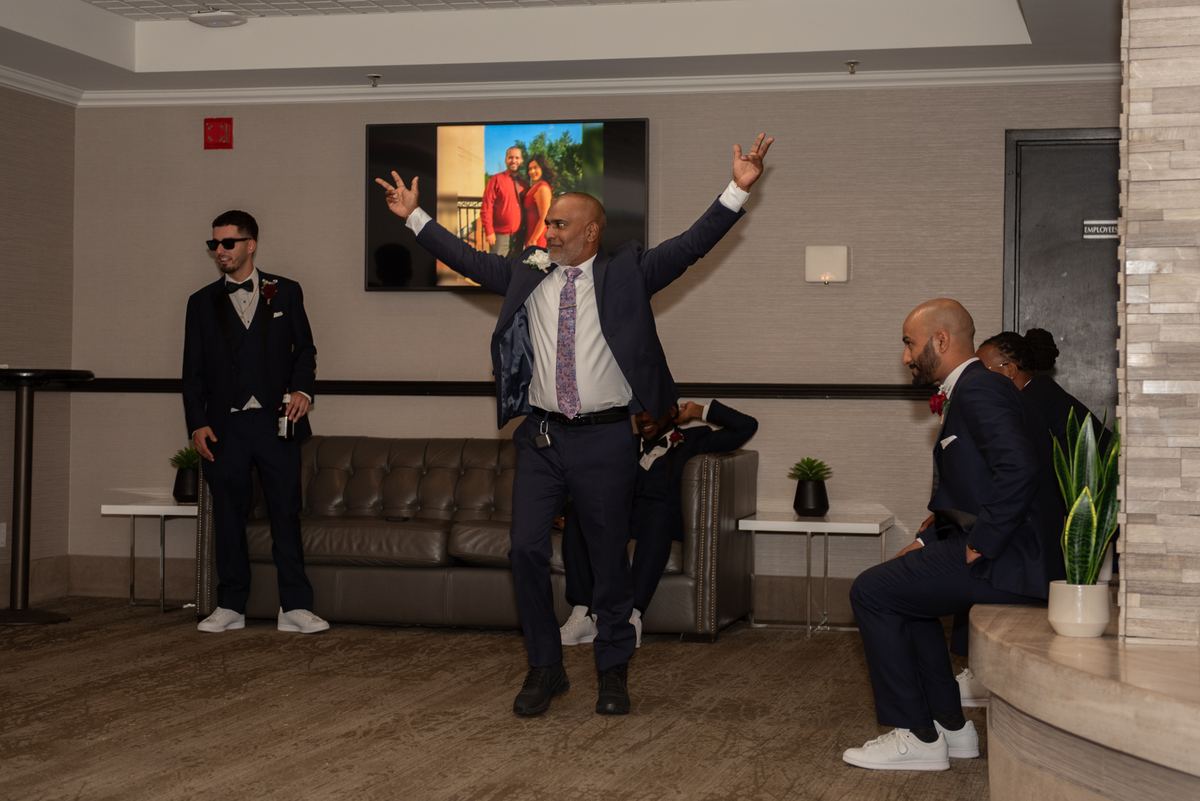 A man in a suit dances with raised arms, surrounded by amused groomsmen in the lounge, near a TV showing a couple's photo.