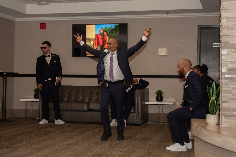 A man in a suit dances with raised arms, surrounded by amused groomsmen in the lounge, near a TV showing a couple's photo.