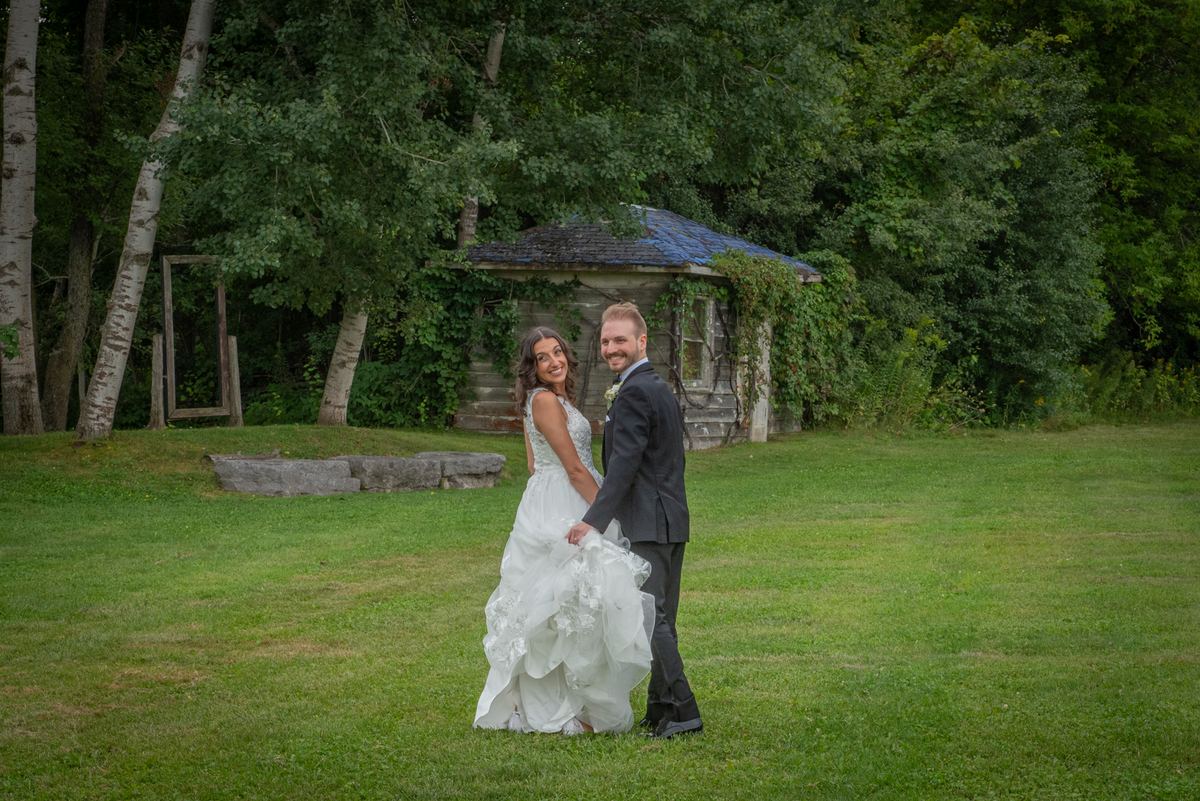 A smiling bride and groom stand hand in hand on a grassy lawn, with a rustic stone structure and birch trees behind them.