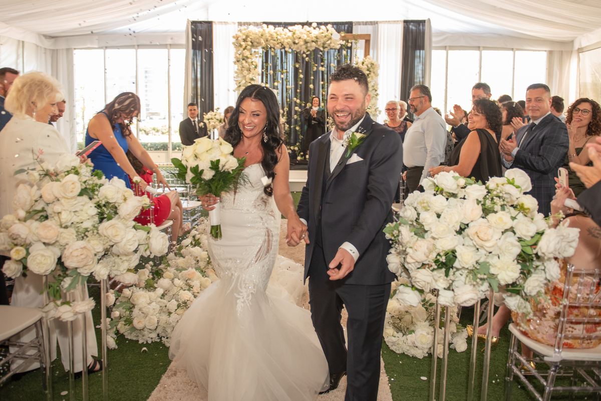 A smiling bride and groom walk down a flower-lined aisle under a white tent as guests clap, bathed in natural light.