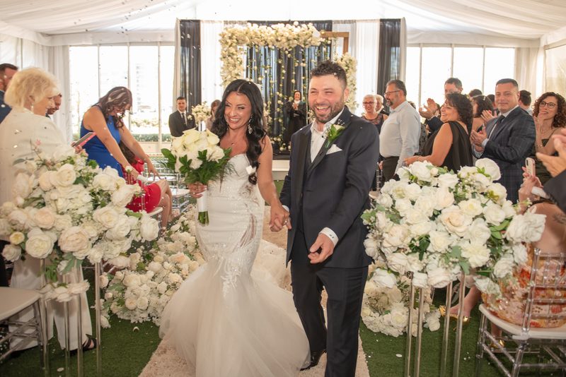 A smiling bride and groom walk down a flower-lined aisle under a white tent as guests clap, bathed in natural light.