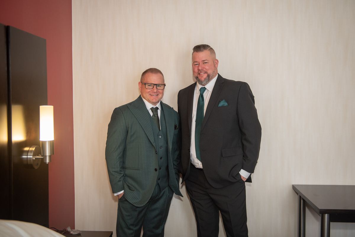 Two men in suits stand casually with hands in pockets near a modern wall sconce in a hotel room, illuminated by soft light.