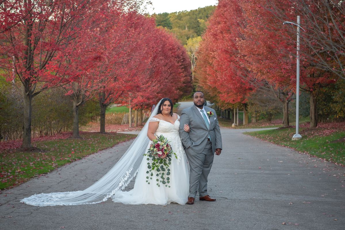 A bride in white and groom in gray stand together, framed by autumn trees along a path bathed in warm light.