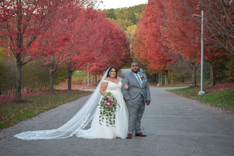A bride in white and groom in gray stand together, framed by autumn trees along a path bathed in warm light.