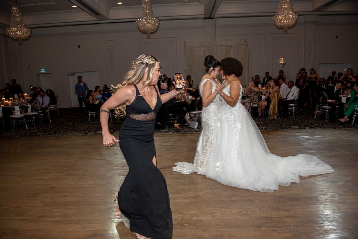 Two brides dance intimately on a polished floor as guests watch and chandeliers glow, celebrating the moment.