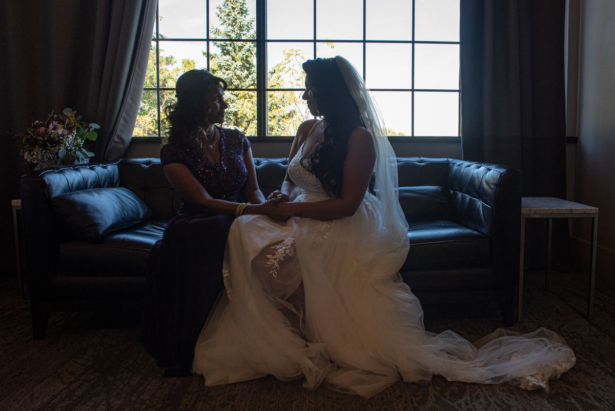 A bride and a woman in an elegant gown sit on a leather couch, holding hands, bathed in warm daylight from a large window.