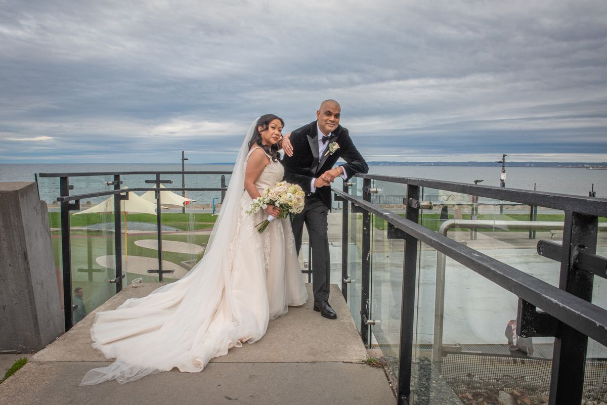 A bride in a white gown and veil leans against her groom in a tuxedo on a waterfront promenade, with the water.