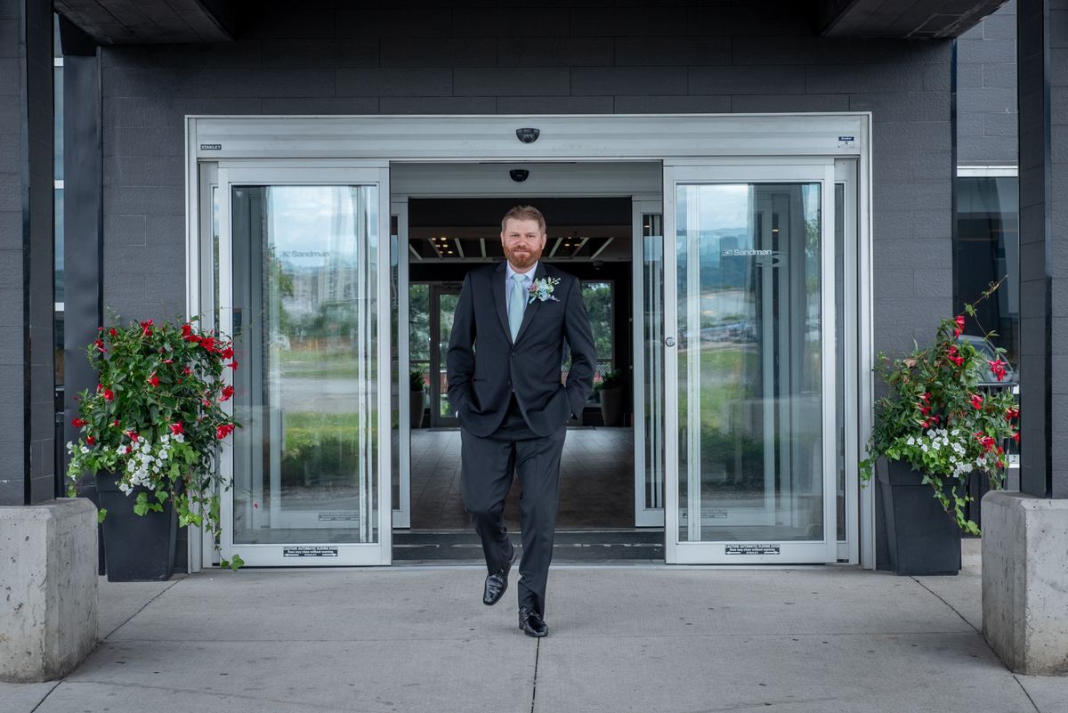 A groom in a dark suit and light blue tie strides confidently through glass doors, hands in pockets, with potted plants.