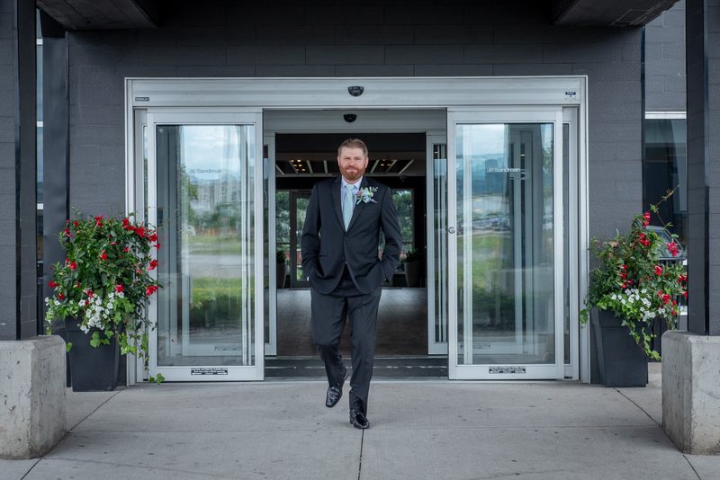 A groom in a dark suit and light blue tie strides confidently through glass doors, hands in pockets, with potted plants.