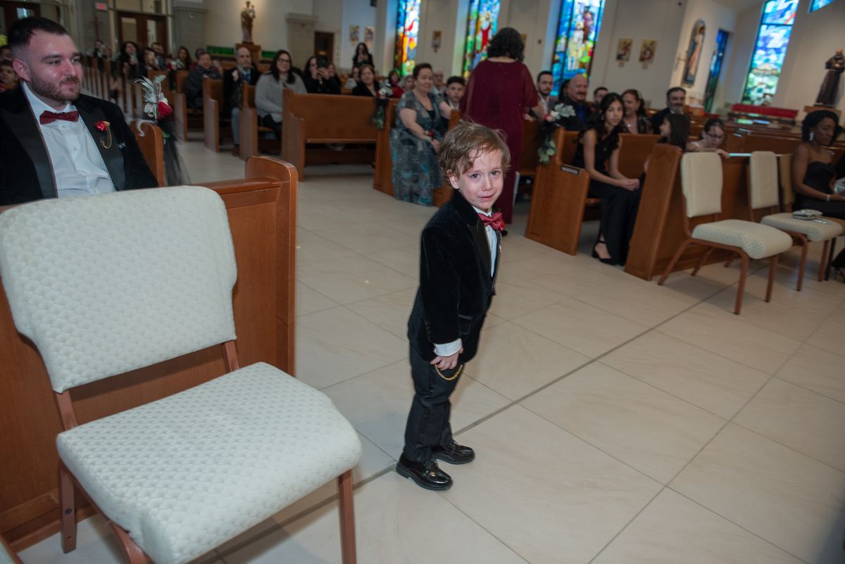 A young boy in a black tuxedo with a red bow tie stands curiously in a church aisle during a wedding.