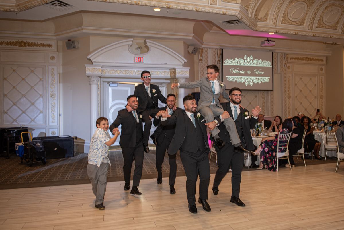 Groomsmen carry the groom across the dance floor as a young boy dances beside them, guests watch with amusement.