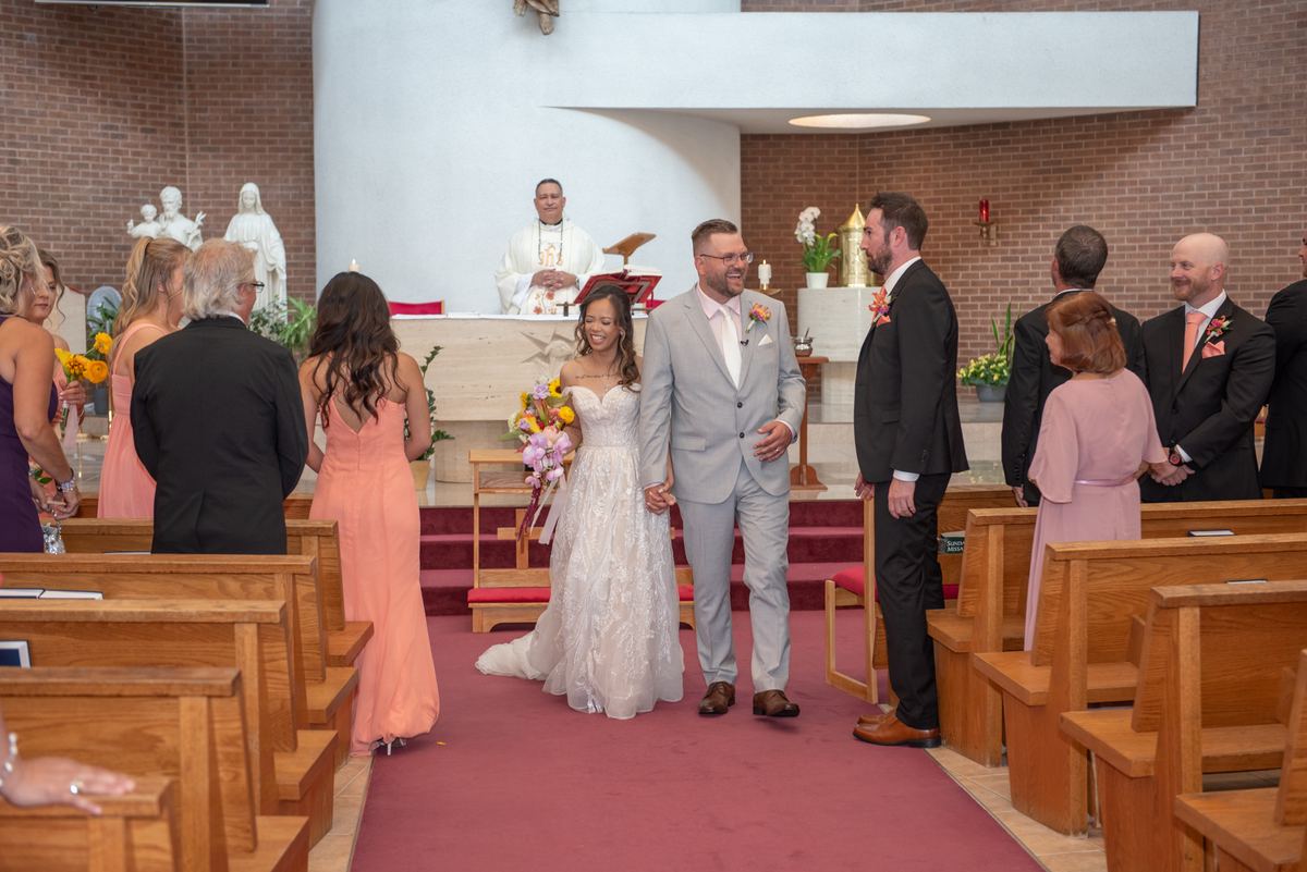 Bride and groom walk down red-carpeted aisle of church, smiling and holding hands, as guests watch from wooden pews.