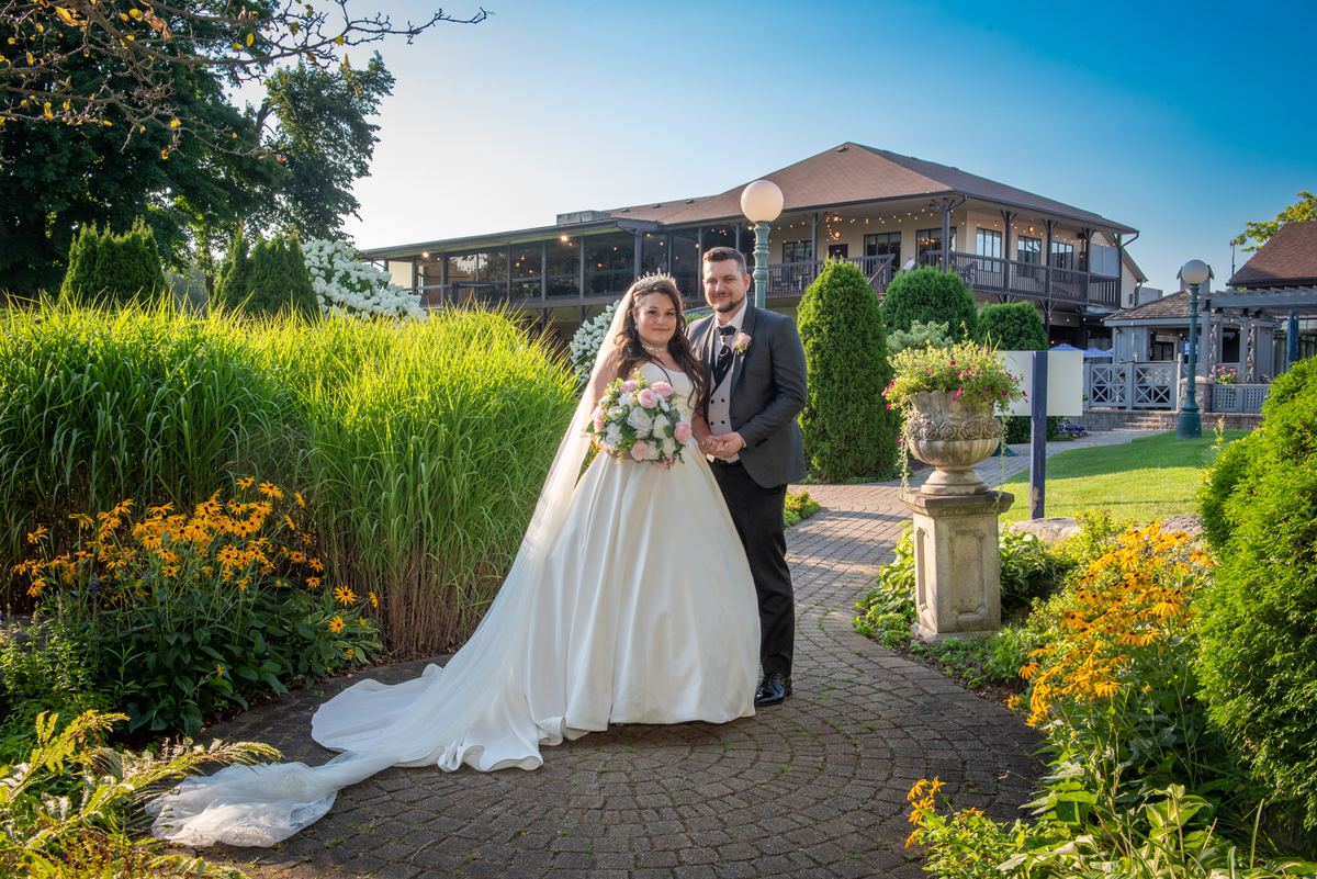 A bride and groom smile on a stone pathway in a lush garden bathed in golden light, surrounded by greenery and flowers.