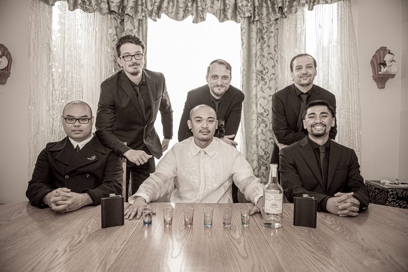 Seven formally dressed men gather around a wooden table with shot glasses, celebrating in diffused light.
