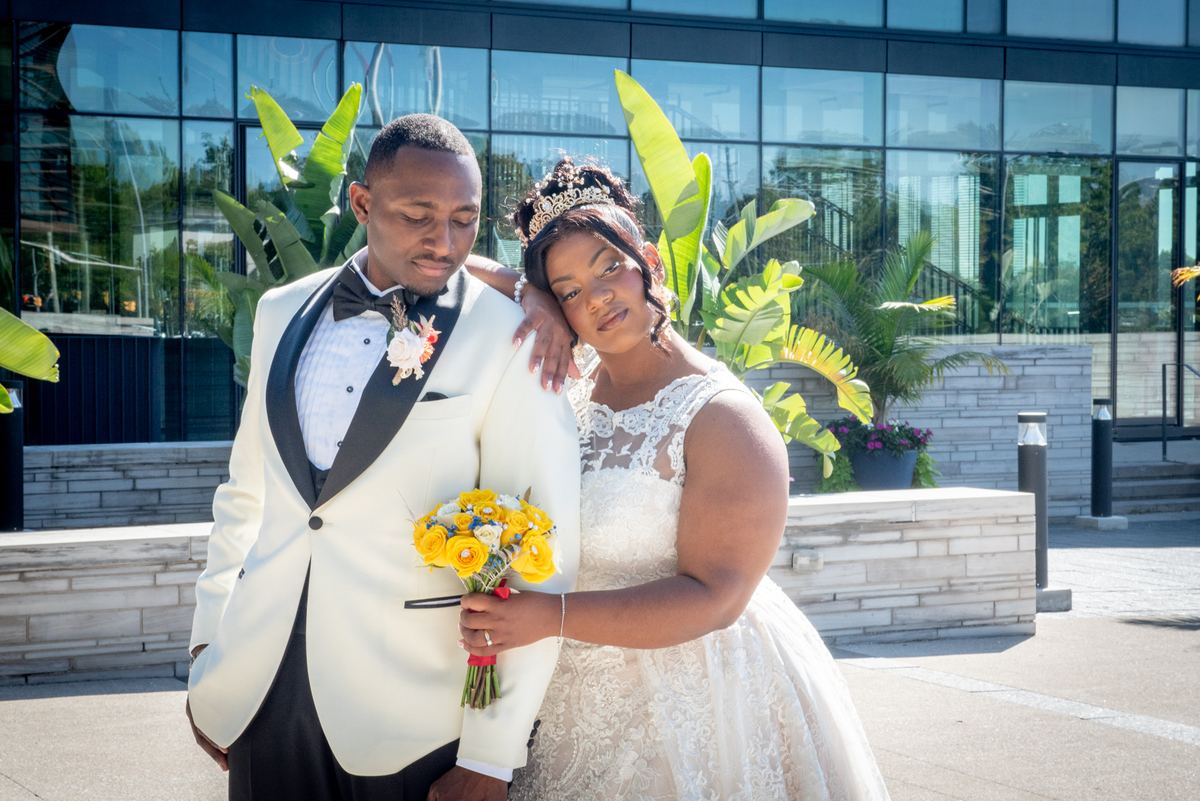 A bride in white lace and groom in tuxedo stand close, gazing at each other by modern glass building and greenery.