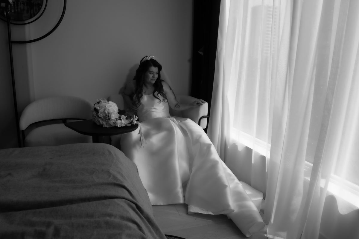 A bride, seated beside a window with sheer curtains in a softly lit room, gazes thoughtfully downward.