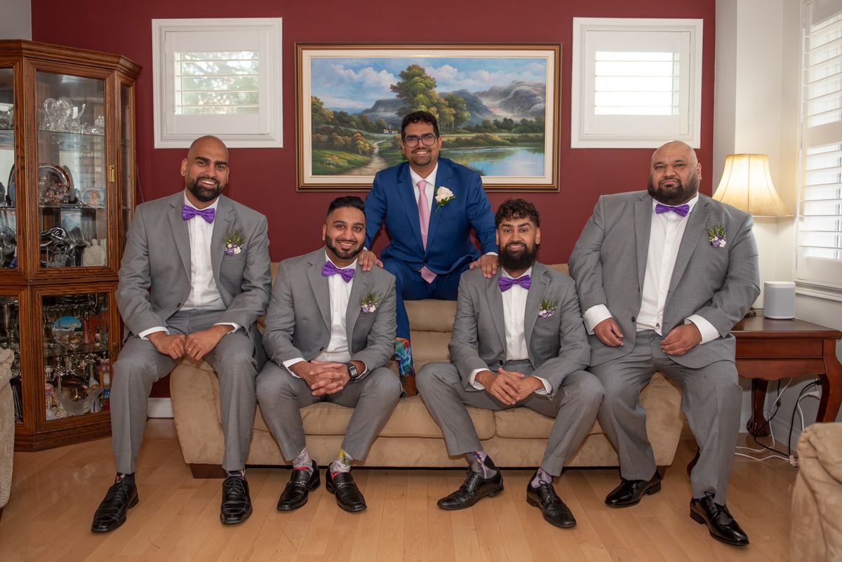 A groom in a blue suit and his four gray-suited, purple-tied groomsmen smile on a beige sofa in a warmly lit living room.