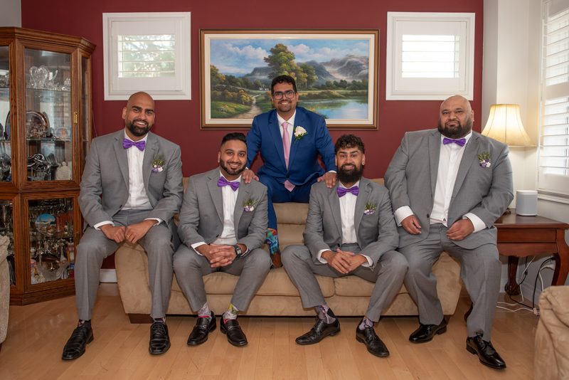 A groom in a blue suit and his four gray-suited, purple-tied groomsmen smile on a beige sofa in a warmly lit living room.