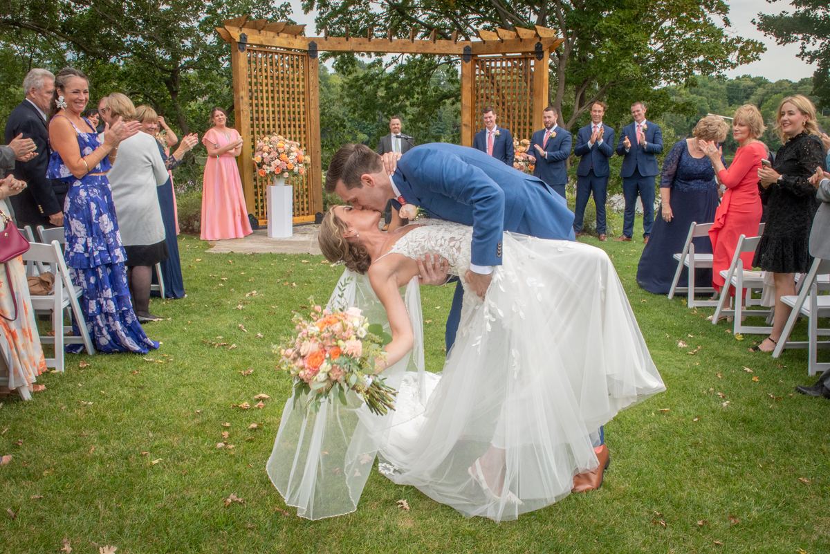 Bride and groom share a playful kiss surrounded by clapping guests in an outdoor ceremony under a wooden arbor.