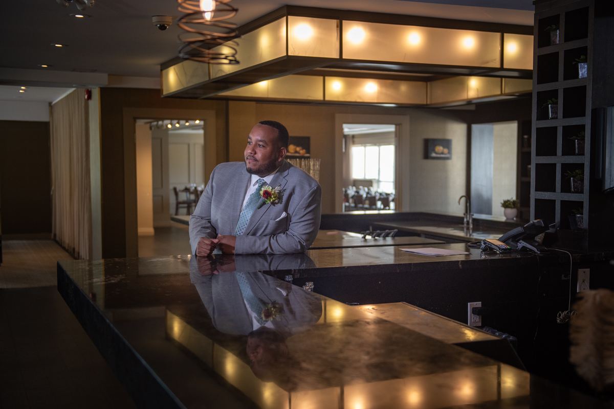 A groom in a gray suit stands thoughtfully beside a glossy black reception desk in a warmly lit hotel lobby.
