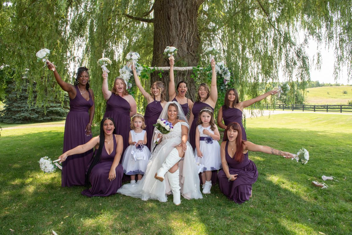 A bride and her bridal party joyfully toss bouquets under a weeping willow, sunlight filtering through the leaves.