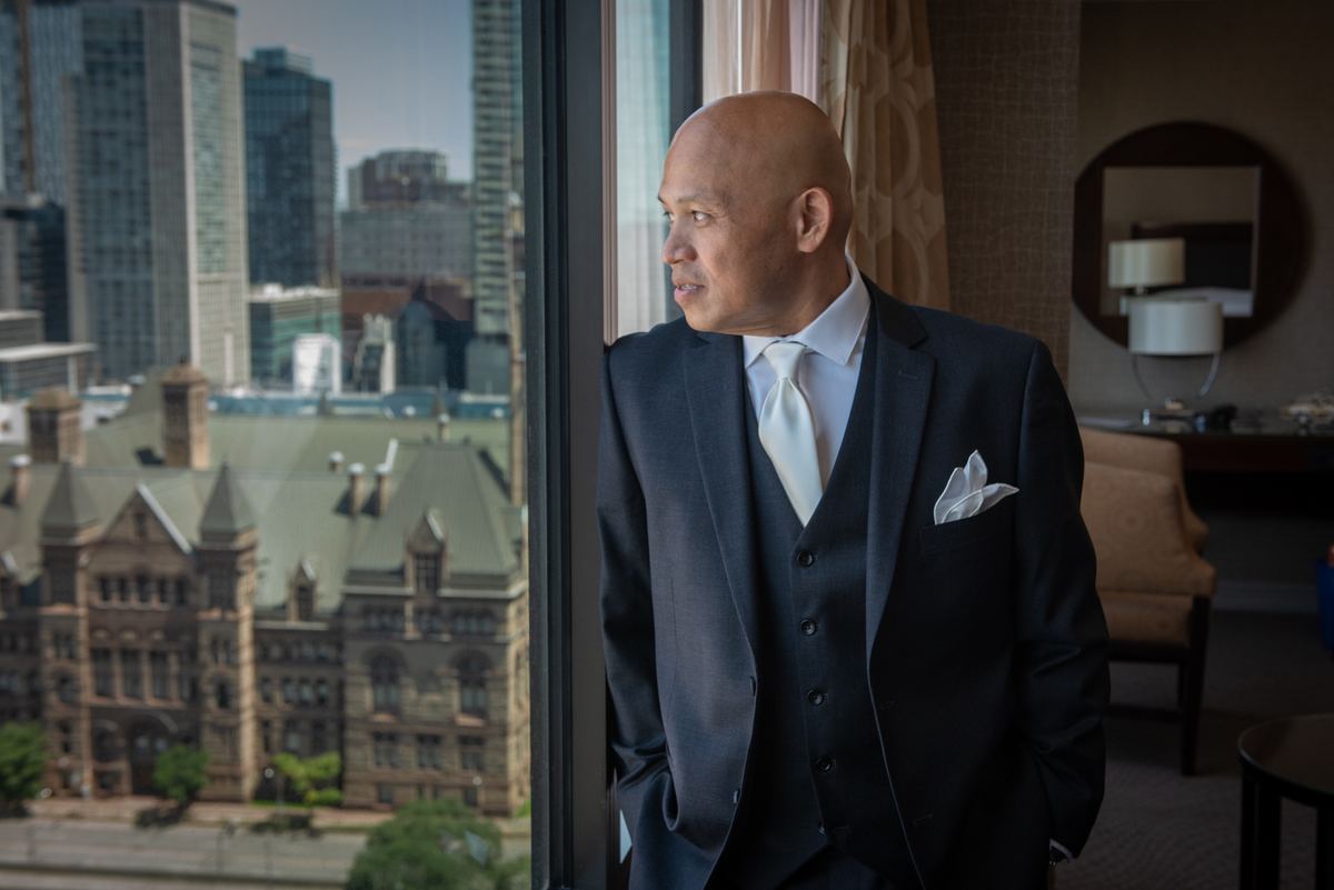 A man in formal wear stands by a window, contemplating the mix of historic and modern architecture outside.