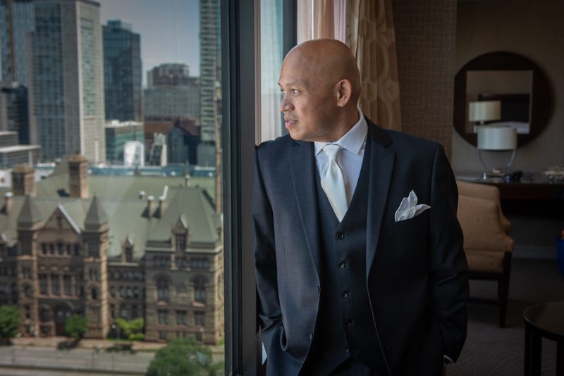 A man in formal wear stands by a window, contemplating the mix of historic and modern architecture outside.