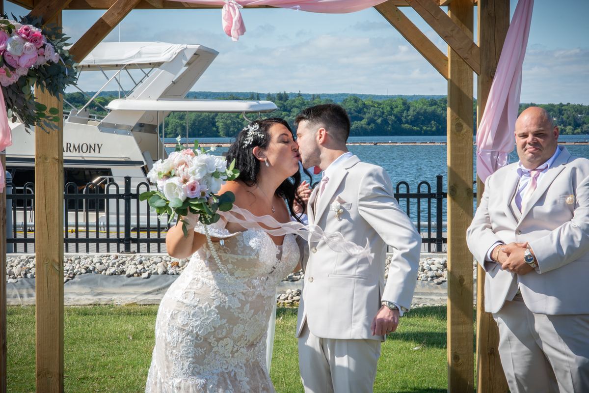 A bride and groom kiss beneath a wooden arch trimmed with pink ribbons and flowers, a yacht on the sunlit water behind them.