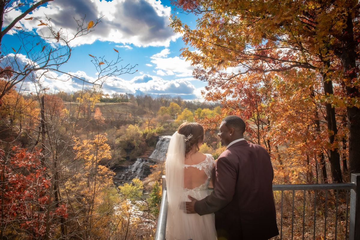 A newlywed couple stands on a platform, gazing at a scenic autumn landscape with a waterfall and colorful foliage.