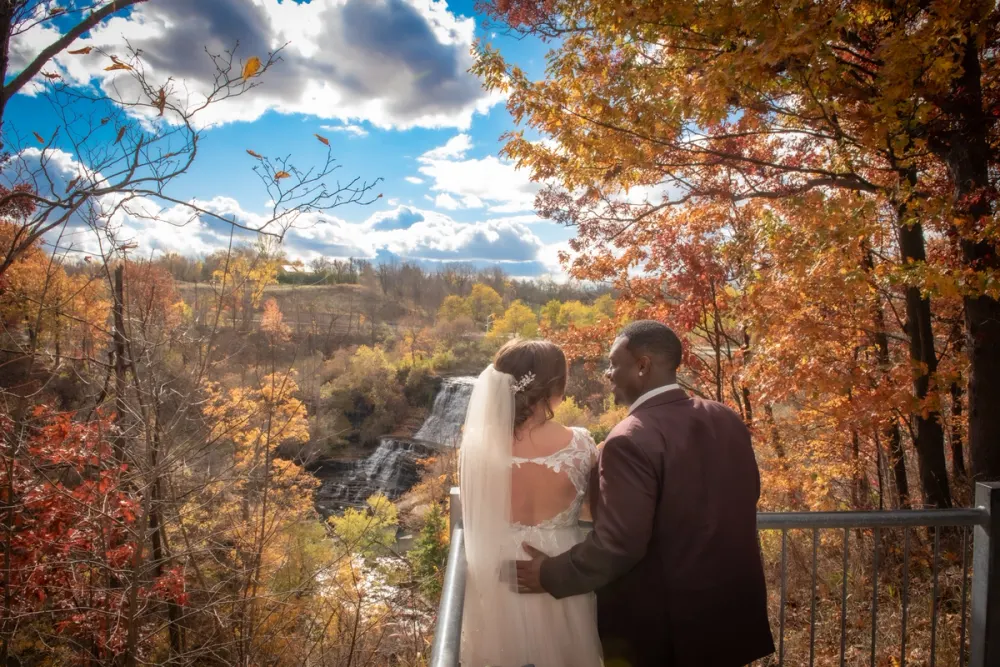 A newlywed couple stands on a platform, gazing at a scenic autumn landscape with a waterfall and colorful foliage.