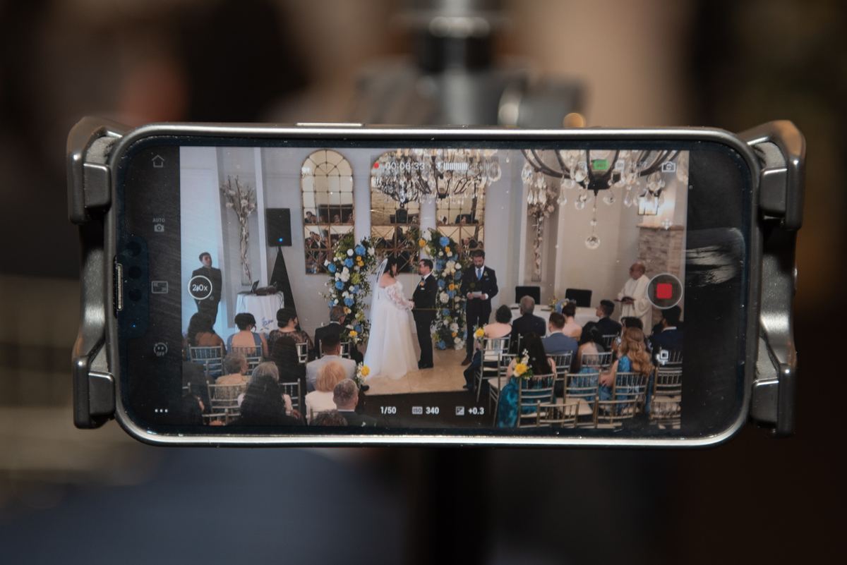 Bride and groom stand at an altar with blue and white flowers, guests seated around them in soft light from chandeliers.