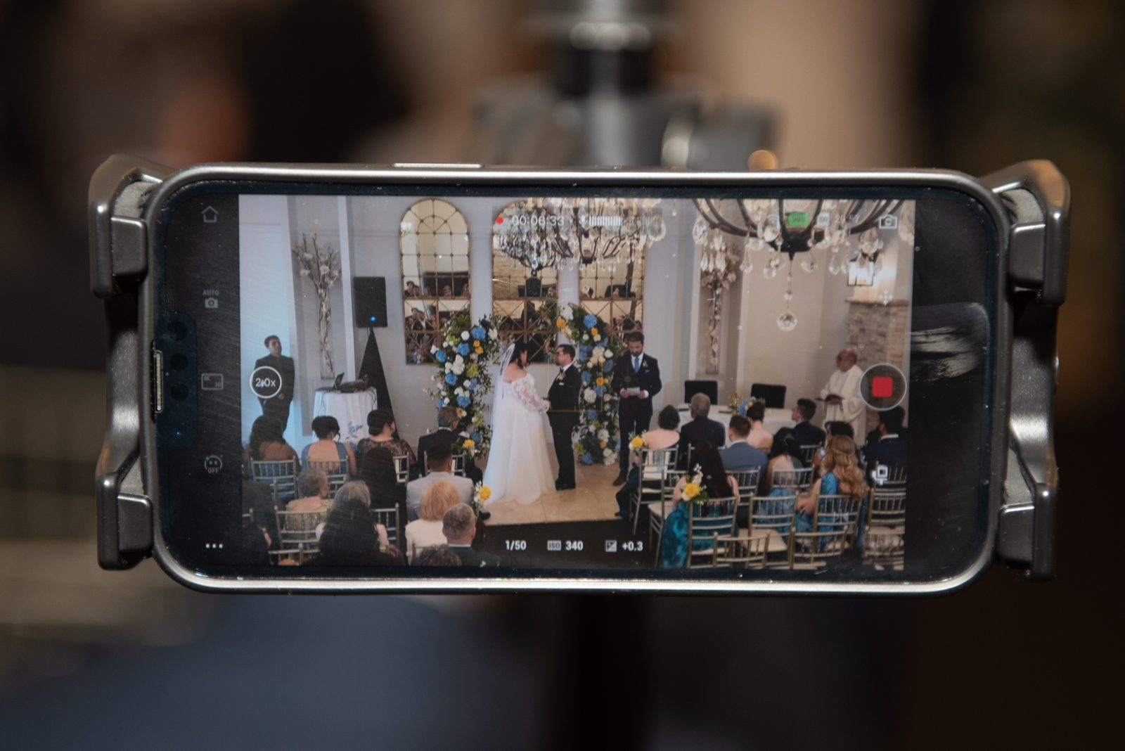 Bride and groom stand at an altar with blue and white flowers, guests seated around them in soft light from chandeliers.