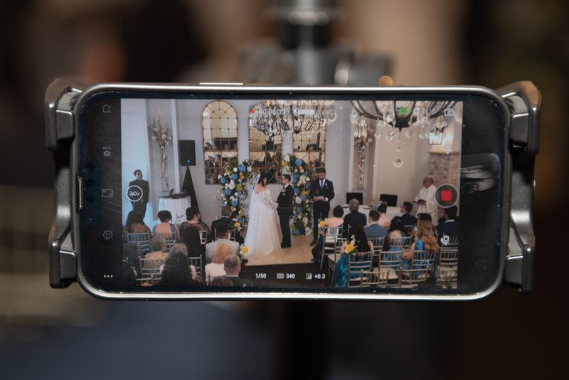 Bride and groom stand at an altar with blue and white flowers, guests seated around them in soft light from chandeliers.