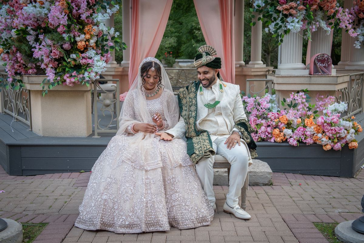 A South Asian bride and groom in traditional attire smile on a garden pavilion bench surrounded by floral arrangements.