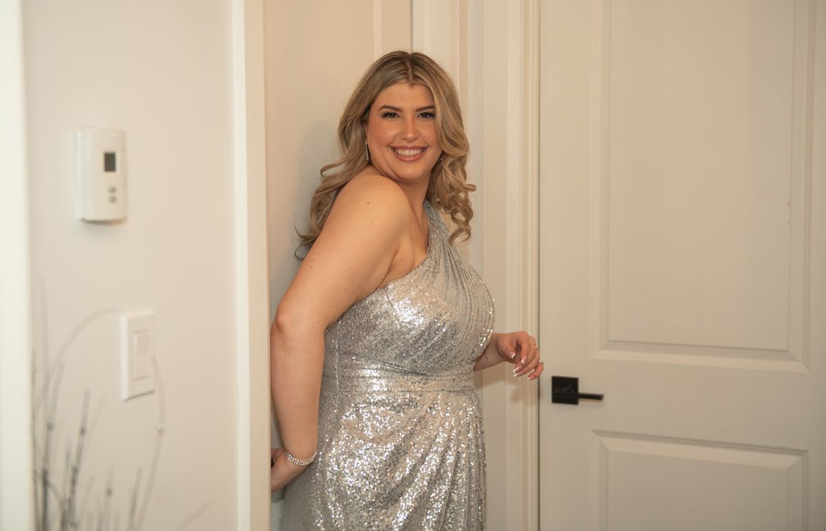A woman in a silver sequined gown smiles joyfully from behind a doorway, her hand resting on the frame.
