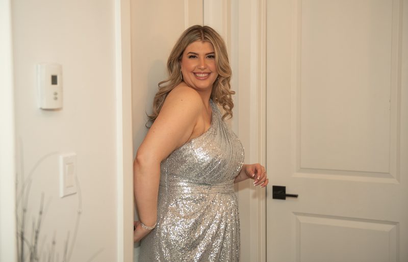 A woman in a silver sequined gown smiles joyfully from behind a doorway, her hand resting on the frame.