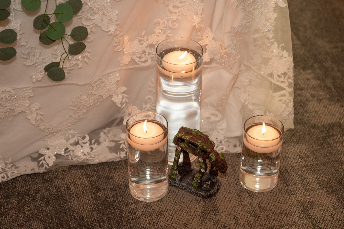 Three candles with a lace curtain and eucalyptus sprig beside a moss-covered AT-AT figurine create an intimate scene.