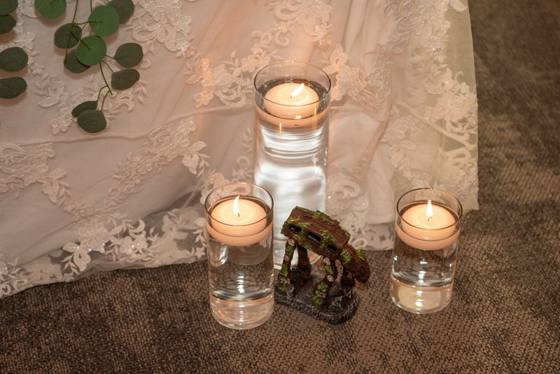 Three candles with a lace curtain and eucalyptus sprig beside a moss-covered AT-AT figurine create an intimate scene.
