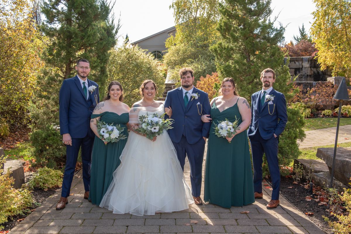 A joyful wedding party poses outdoors on a paved path surrounded by autumn foliage and trees, smiling at the camera.