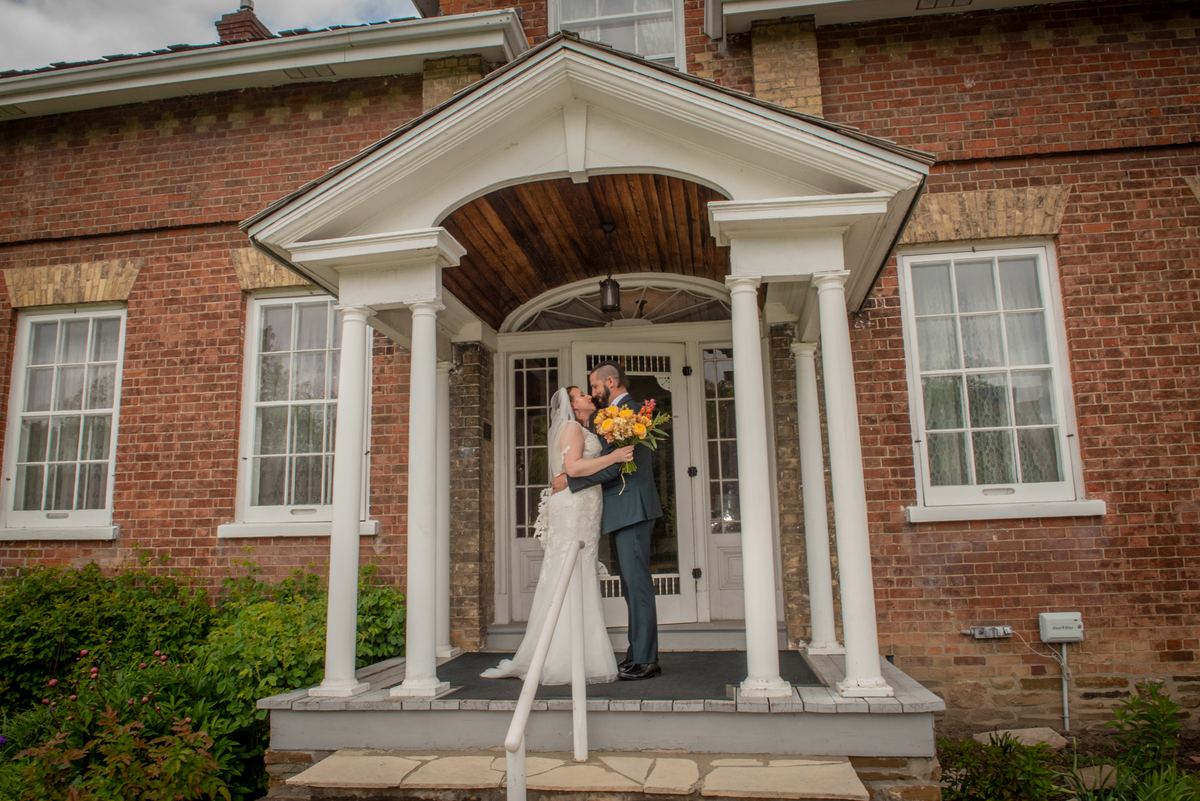 Jenna-Marie and Michael share a tender kiss on the porch of a red brick house, framed by white columns and a wooden portico.