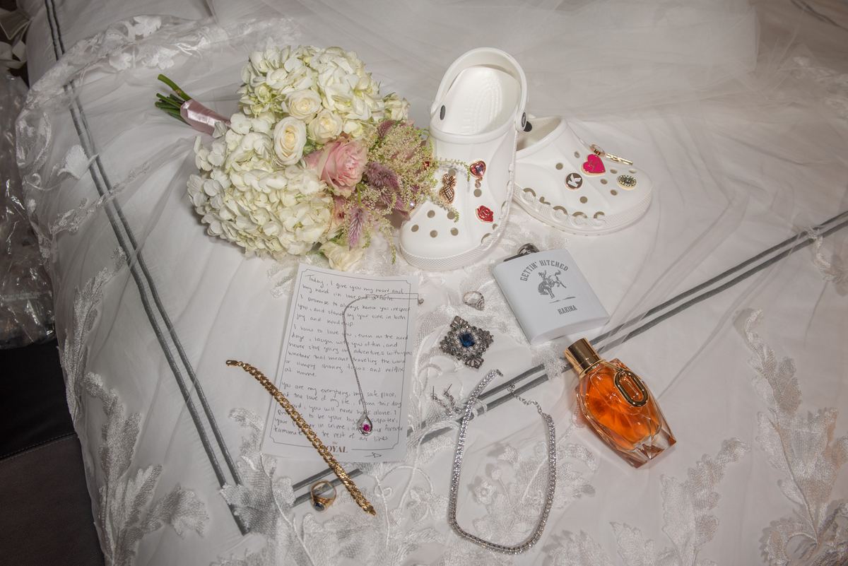 A bed with a bouquet of white and pink flowers, Crocs, vows, jewelry, perfume, and flask for a wedding.