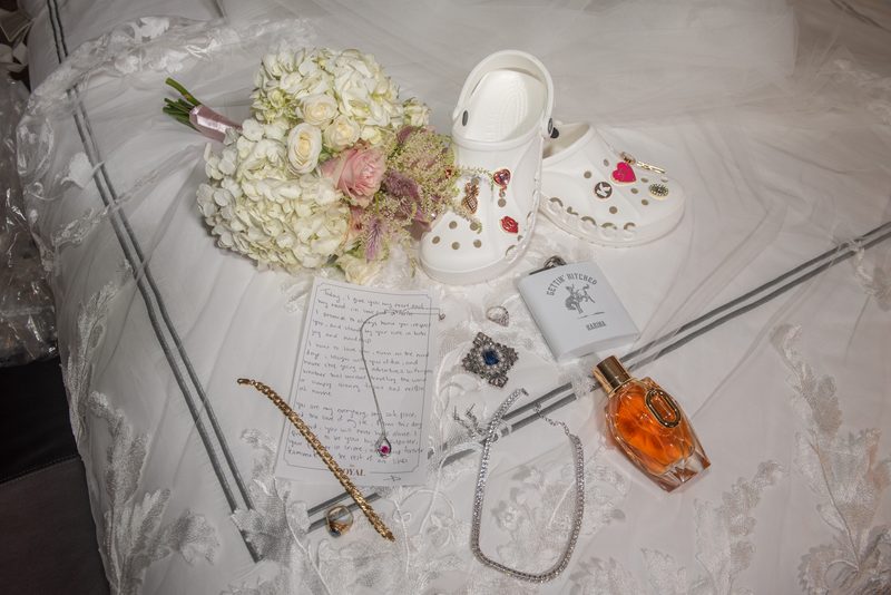 A bed with a bouquet of white and pink flowers, Crocs, vows, jewelry, perfume, and flask for a wedding.