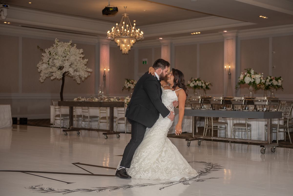 A newlywed couple dances joyfully on a polished ballroom floor, surrounded by elegant decor and warm lighting.