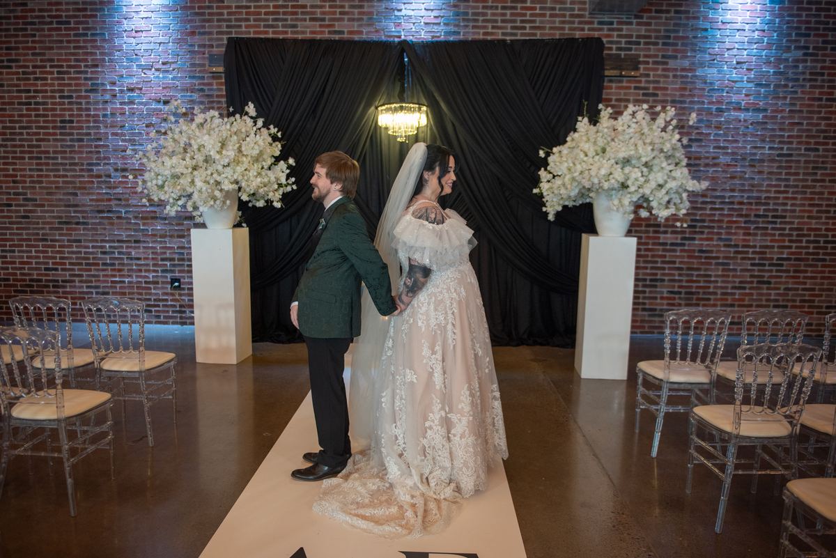 Bride and groom stand on white runner, smiling at each other near black-draped altar with floral arrangements.