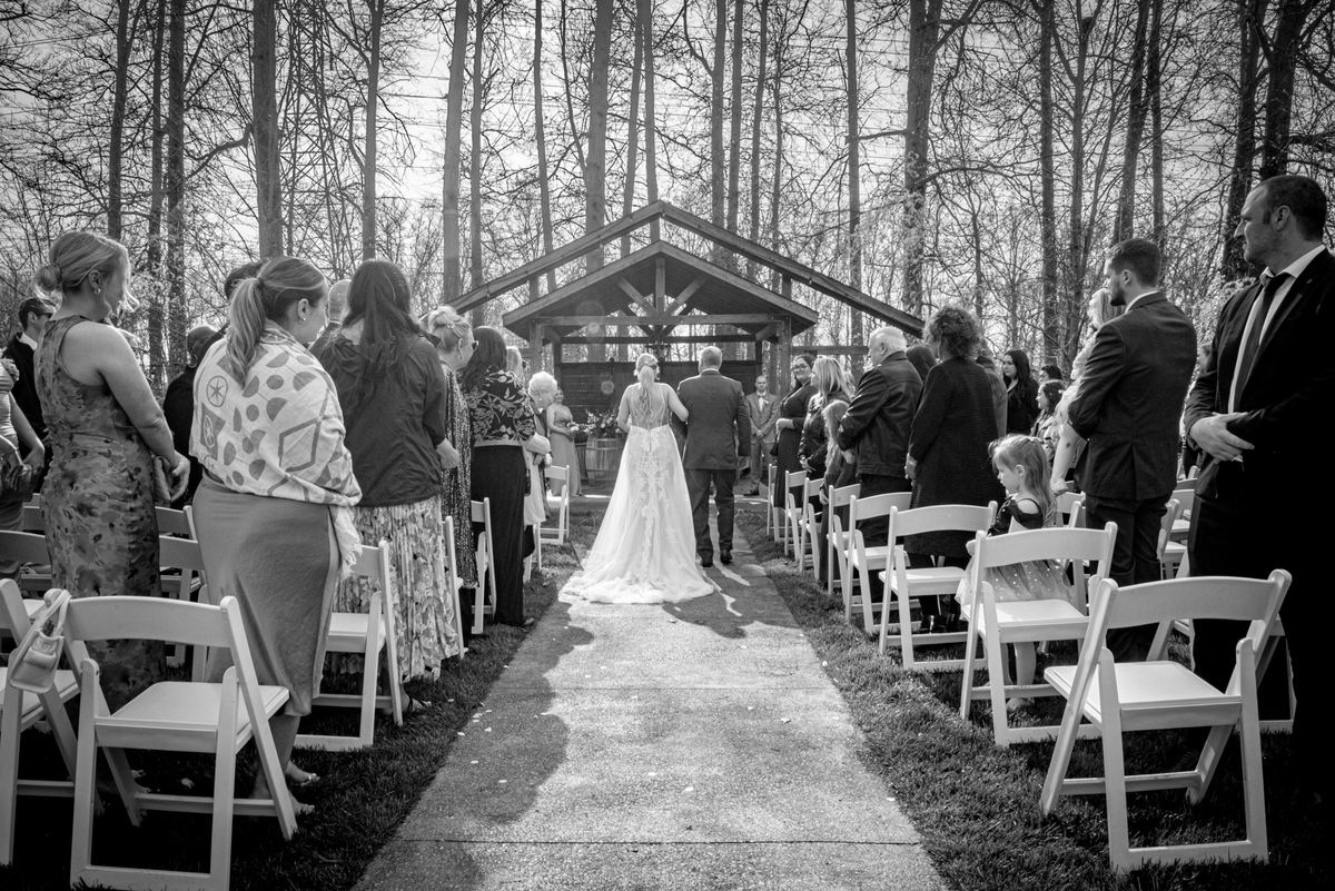 A bride walks down an aisle toward a wooden arbor, escorted by a man, as guests watch in a sun-dappled wooded clearing.