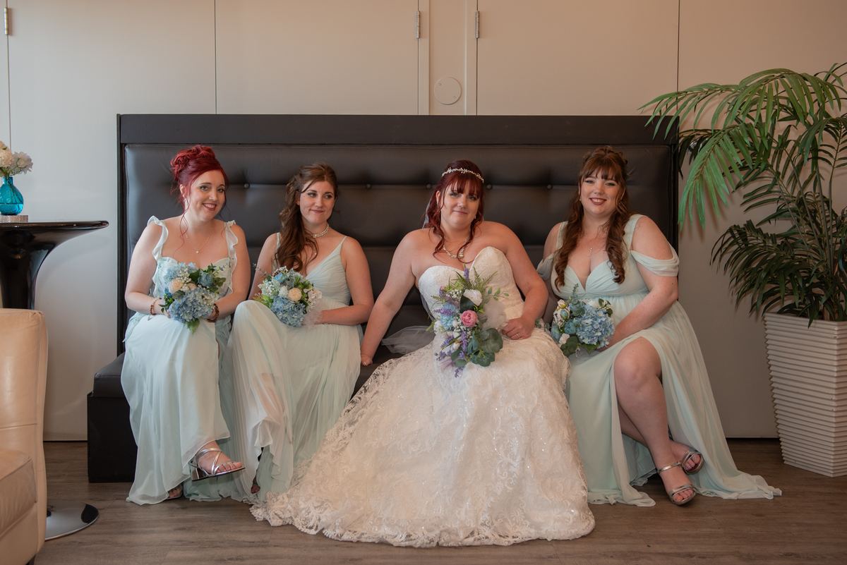 Four women, including the bride in a white lace gown, sit on a dark upholstered bench holding blue and white flower bouquets.